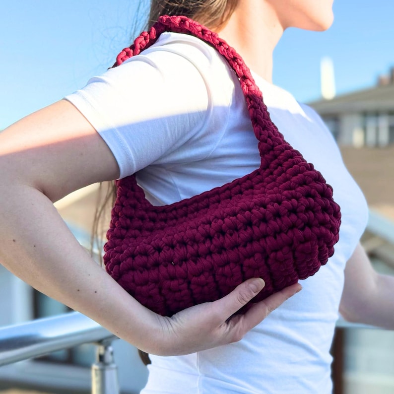 May include: A burgundy crochet shoulder bag with a thick strap. The bag is held by a person wearing a white t-shirt. The bag's texture is visible, and the background is blurred.