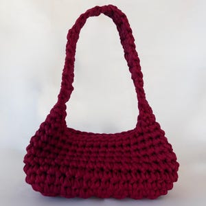 May include: A burgundy crochet handbag with a rounded base and braided handles. The bag is made of thick yarn, giving it a textured appearance. The handles curve upwards, creating a stylish, shoulder bag design.
