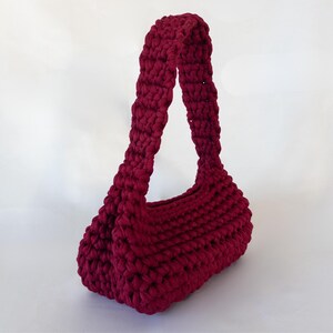 May include: A burgundy crochet handbag with a thick, textured stitch. The bag features a rounded base and a single, wide handle. The color is a deep, rich red, and the bag appears to be handmade.