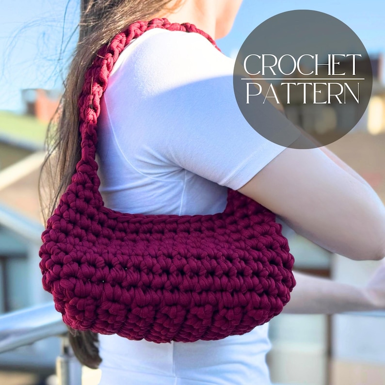 May include: A burgundy crochet shoulder bag with a rounded shape. The bag is made of thick yarn and has a matching strap. The image includes a circular graphic with the words "CROCHET PATTERN".