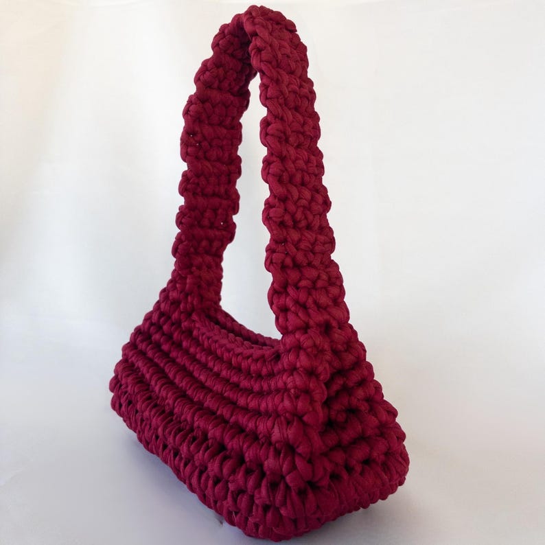May include: A burgundy-colored, hand-crocheted handbag with a rectangular base and a curved handle. The bag's body features horizontal ribbing, and the handle is thick and textured. The bag is made of a soft, chunky yarn.