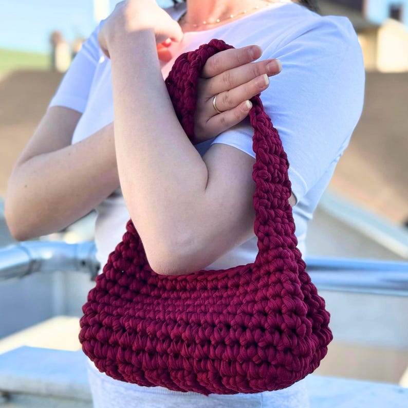 May include: A burgundy crochet handbag with a rounded shape and a thick, textured strap. The bag is held by a person wearing a white t-shirt. The bag's color is a deep, rich shade of red.