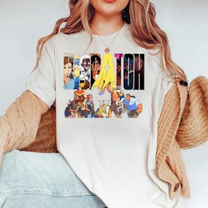 May include: A cream-coloured t-shirt with the word "DEPATOH" in a stylised font, featuring a superhero theme. The design includes various superhero characters and a yellow cape. The shirt is made of a soft material.