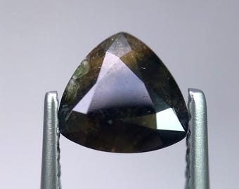 May include: A triangular-cut gemstone held by metal tweezers. The gem has a dark, olive-green hue with hints of purple and brown. The facets reflect light, creating a sparkling effect. The background is a soft, neutral gray.