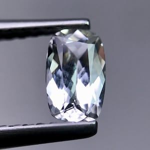 May include: A faceted, light-colored gemstone held by a pair of tweezers. The gem has a rectangular cushion cut with many reflective surfaces. The gem's color is a pale, translucent blue-green, and it is set against a gray background.