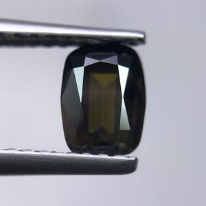 May include: A faceted, olive-green gemstone held by tweezers. The cushion-cut stone has a rectangular shape with rounded corners and multiple reflective surfaces. The gem is a deep, earthy green color.