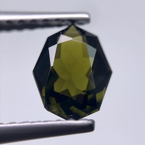May include: A faceted, olive-green gemstone held by a pair of silver tweezers. The gem has a complex, geometric cut, reflecting light and showcasing its clarity. The gemstone's color ranges from a deep, forest green to a lighter, yellowish-green.