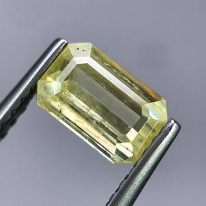 May include: A rectangular, emerald-cut yellow gemstone with a transparent, faceted surface. The stone is held by a pair of silver-toned metal tweezers, showcasing its clear, pale yellow color and geometric cut against a neutral background.