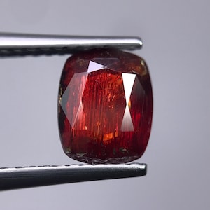 Puede incluir: Una piedra preciosa de talla cojín con un color rojo intenso e inclusiones internas visibles. La piedra presenta una superficie facetada que refleja la luz, mostrando diversos tonos de carmesí y naranja en su estructura translúcida.