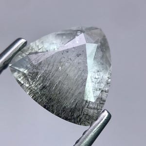 Può includere: Una gemma triangolare sfaccettata con inclusioni interne, tenuta da pinzette metalliche. La gemma è principalmente trasparente con aree nere e grigie, creando un aspetto strutturato. La forma della gemma è un triangolo.