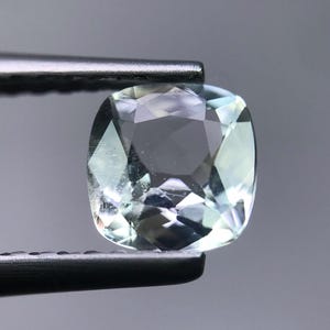 May include: A faceted, cushion-cut gemstone held by tweezers. The gem has a pale green and blue color with internal facets that reflect light. The tweezers are silver-colored and hold the gem against a gray background.