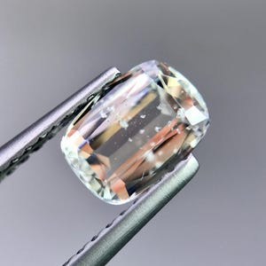May include: A faceted, rectangular gemstone held by tweezers. The gem is primarily clear with internal inclusions and subtle peach and yellow hues. The facets reflect light, creating a sparkling effect. The background is a soft gray.