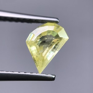 May include: A faceted, shield-shaped yellow gemstone with a translucent appearance and sharp, angular edges. The stone displays a pale lemon-yellow hue with internal light reflections and visible facets across its surface, held by metal tweezers.