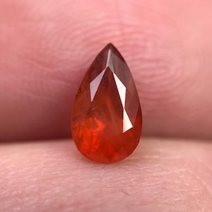 以下が含まれることがあります： 鮮やかなオレンジレッド色の、ファセット加工された涙型の宝石。宝石の表面は光を反射し、その多面的なカットと内部の透明度を示しています。宝石は柔らかくニュートラルな背景に配置されています。