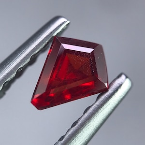 May include: A faceted, kite-shaped, deep red gemstone is held by silver tweezers against a grey background. The gem has sharp, clean edges and reflects light, showcasing its internal structure. The tweezers provide a contrasting metallic element.