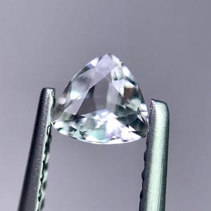 May include: A clear, triangular gemstone held by silver tweezers. The gem has multiple facets that reflect light, creating a sparkling effect. The background is a soft gray, highlighting the gem's clarity and shape. This image showcases a precious stone.