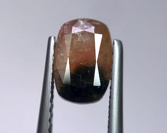 May include: A faceted gemstone held by tweezers. The gem has a mix of colors, including pink, brown, and black. The gem is oval-shaped with multiple facets that reflect light. The tweezers are silver-colored.
