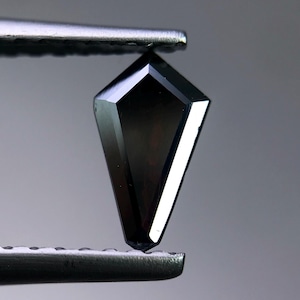 Puede incluir: Una gema negra facetada en forma de cometa es sostenida por unas pinzas. La gema tiene una superficie pulida con múltiples facetas que reflejan la luz. El color oscuro y la forma única de la gema la convierten en una pieza llamativa.