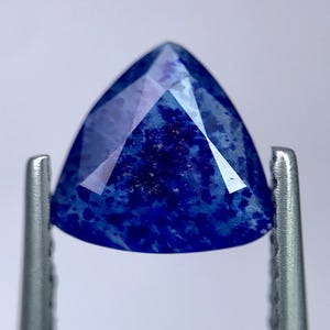 Peut inclure: Une pierre précieuse triangulaire et facettée, d'un bleu profond, tenue par une pince argentée. La surface de la gemme reflète la lumière, mettant en valeur sa taille et ses motifs internes. La couleur de la pierre est un bleu riche et saturé.