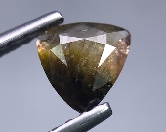 May include: A faceted, triangular gemstone held by silver tweezers. The gem has a mix of brown, olive, and clear tones, with sharp angles and a polished surface. The gem is set against a soft gray background.