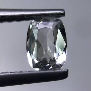 May include: A faceted, light green gemstone held by a pair of tweezers. The gem has a rectangular cushion cut with multiple facets that reflect light. The stone appears to be transparent with a slight green hue.