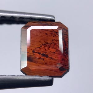 Puede incluir: Una gema de talla esmeralda sujeta con pinzas. La gema presenta un tono marrón rojizo cálido con inclusiones oscuras y una superficie facetada reflectante. Los bordes están bien definidos, mostrando la forma de la gema.