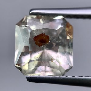 May include: A clear, emerald-cut gemstone with internal inclusions, held by a pair of tweezers. The gem has a light, almost colorless appearance with a few small, reddish-brown inclusions visible within the facets.