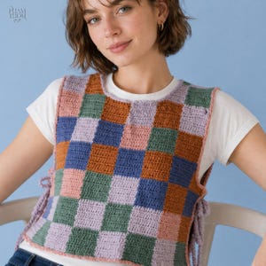 May include: A colorful, crochet vest with a checkerboard pattern in shades of pink, green, blue, and brown. The vest has side ties and is worn over a white t-shirt. The background is a solid light blue.