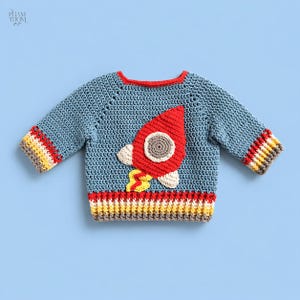 May include: A blue crochet sweater with a red rocket ship applique. The rocket has a gray circle and off-white accents. The cuffs and bottom hem feature stripes of red, yellow, and brown. The sweater is designed for a child.