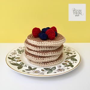 Könnte beinhalten: Ein Stapel gehäkelter Pfannkuchen mit roten und blauen Beerenbelägen, präsentiert auf einem Teller mit floralem Muster. Die Pfannkuchen sind beige und braun, und der Teller hat einen goldenen Rand. Der Hintergrund ist einfarbig gelb.