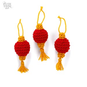 Könnte beinhalten: Drei handgefertigte rote und gelbe Häkellaternen. Jede Laterne hat einen runden roten Körper, einen gelben oberen Teil, eine gelbe Quaste und eine gelbe Schlaufe zum Aufhängen. Die Laternen werden vor einem weißen Hintergrund präsentiert.