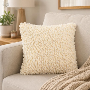 May include: A square, cream-colored decorative pillow with a textured, looped surface. The pillow is placed on a light beige sofa, with a tan throw blanket visible in the corner. The pillow adds a cozy element to the living room.