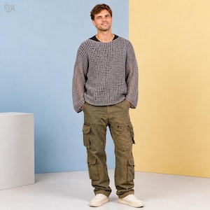 Op de afbeelding: Een persoon draagt een grijze, open geweven trui over een zwart shirt, gecombineerd met olijfgroene cargobroeken en witte sneakers. De achtergrond is verdeeld in blauwe en gele secties.