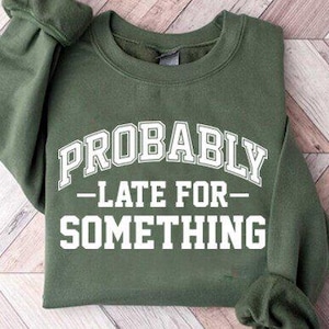 May include: Olive green sweatshirt with the text "PROBABLY - LATE FOR - SOMETHING" in white, arched lettering. The sweatshirt is folded on a light brown and white chevron patterned surface.