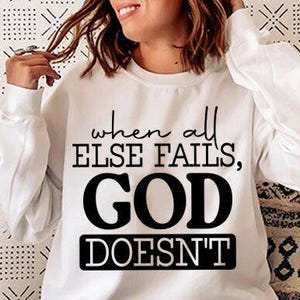 May include: A white sweatshirt featuring the black text "when all else fails, GOD DOESN'T". The long-sleeved top has the text centered. The image includes a person and a patterned background.