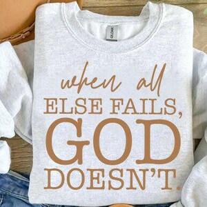 May include: A pale grey sweatshirt featuring the brown text: "when all else fails, GOD doesn't". The sleeves are visible, and the image includes a hat and a pair of jeans. The sweatshirt is folded.