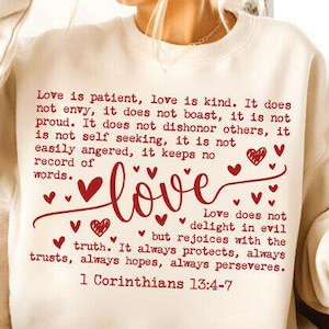 Op de afbeelding: Crèmekleurige sweatshirt met het woord "love" in een sierlijk lettertype, versierd met rode harten en tekst uit 1 Korinthe 13:4-7. Het ontwerp is gecentreerd op de voorkant.