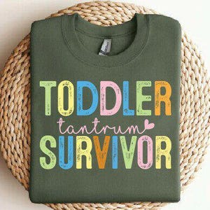May include: Olive green crew neck sweatshirt with the words "TODDLER tantrum SURVIVOR" in colorful block letters. The sweatshirt is displayed on a woven, circular placemat. The text includes a small pink heart.