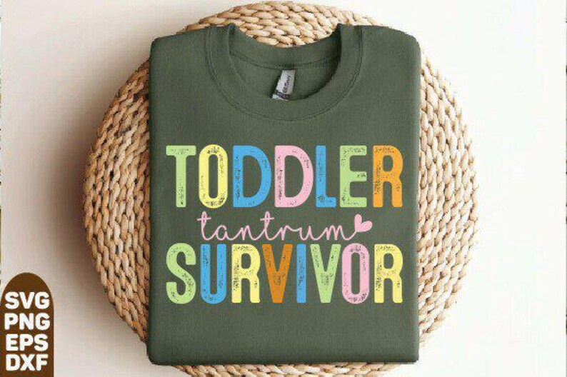 May include: Olive green crew neck sweatshirt with the words "TODDLER tantrum SURVIVOR" in colorful block letters. The sweatshirt is displayed on a woven, circular placemat. The text includes a small pink heart.