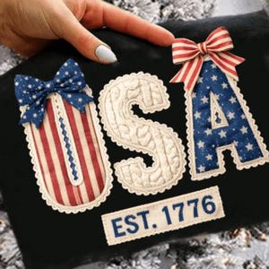 Könnte beinhalten: Schwarzer Stoff mit den Buchstaben "USA" in patriotischen Farben. Das "U" und das "A" sind mit Schleifen verziert. Das "U" hat rot-weiße Streifen, das "A" blaue Sterne und das "S" ist cremefarben. Der Text "EST. 1776" ist ebenfalls vorhanden.