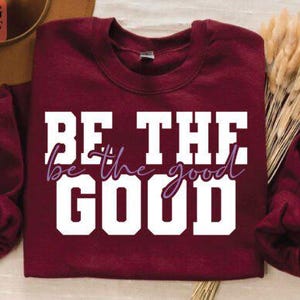 Pode incluir: Uma camisola bordô com as palavras "BE THE GOOD" em letras brancas e cursivas. A camisola está dobrada, com um feixe de espigas de trigo secas e um chapéu marrom ao fundo.