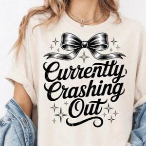 以下が含まれることがあります： 黒いリボンと「Currently Crashing Out」の文字がスタイリッシュなフォントでプリントされた、クリーム色のTシャツ。デザインにはスターバーストのアクセントが含まれています。このシャツはカジュアルで快適なスタイルです。