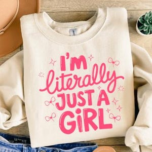 Może przedstawiać: Kremowa bluza z napisem "I'm literally just a girl" w jaskrawym różu. Projekt zawiera gwiazdki i kokardy. Bluza jest połączona z niebieskimi szortami dżinsowymi i brązowym kapeluszem.