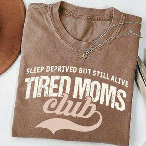 May include: A brown t-shirt with the text "SLEEP DEPRIVED BUT STILL ALIVE TIRED MOMS CLUB" in white and light pink. A brown hat and a silver necklace are also visible. The shirt is folded on a white surface.