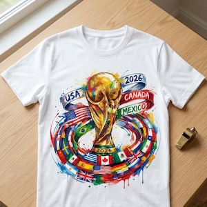 May include: A white t-shirt featuring a central graphic of a gold trophy surrounded by a circular arrangement of various national flags. The design includes the text USA, CANADA, MEXICO, and 2026, accented with colourful watercolour paint splatters.