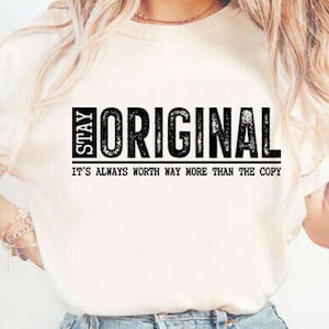 May include: Cream-colored t-shirt with the words "STAY ORIGINAL" in a black, bold font. Below the main text is the phrase "IT'S ALWAYS WORTH WAY MORE THAN THE COPY" in a smaller, black font. The shirt has short sleeves.