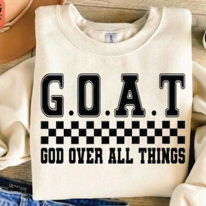 May include: Cream-colored sweatshirt with the text "G.O.A.T. GOD OVER ALL THINGS" in black block letters. A black and white checkered pattern is below the word "G.O.A.T.". The sweatshirt is folded on a wooden surface.