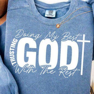 May include: A blue sweatshirt with the text "Doing My Best GOD With The Rest" in white. The word "GOD" is in large capital letters with a cross to the right. A gold necklace with a pendant is visible.