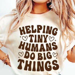 May include: Cream-colored t-shirt with the text "Helping Tiny Humans Do Big Things" in brown, retro-style lettering. The design includes hearts and stars. The shirt is a casual, everyday wear item.
