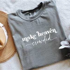 Op de afbeelding: Een grijs t-shirt met de witte tekst "make heaven crowded". Een strooien zonnehoed met een witte band en een takje kunstbloemen liggen naast het shirt. Het shirt is opgevouwen en ligt op een gestructureerd, lichtgekleurd oppervlak.